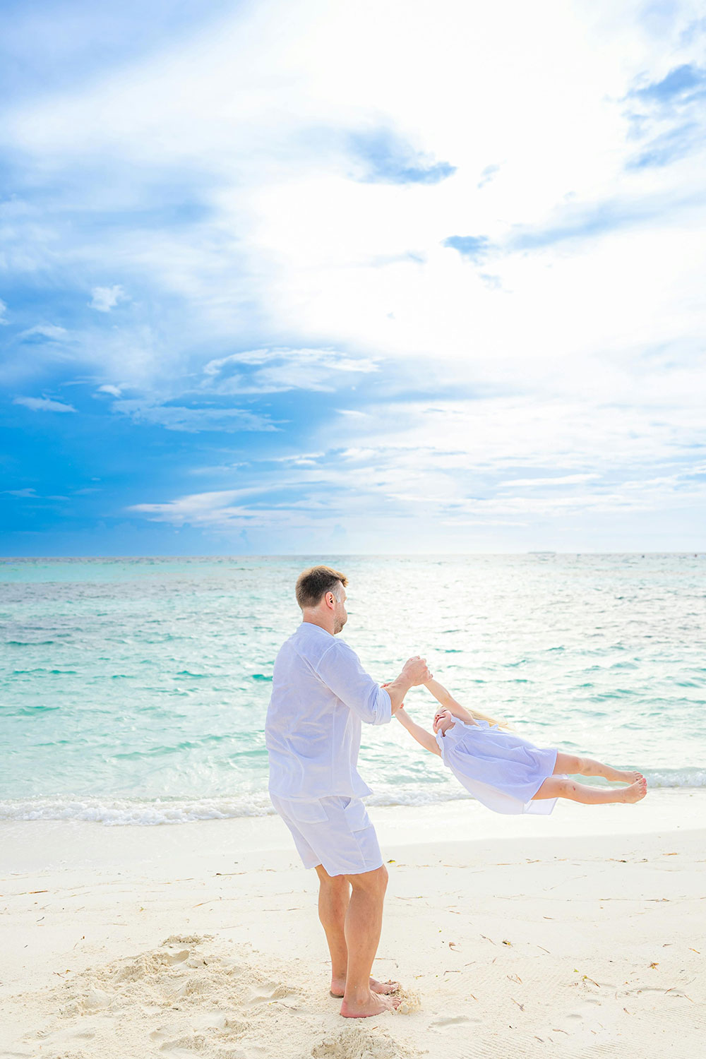 father lifting his young daughter in the air while playing on a white sandy tropical beach near the water