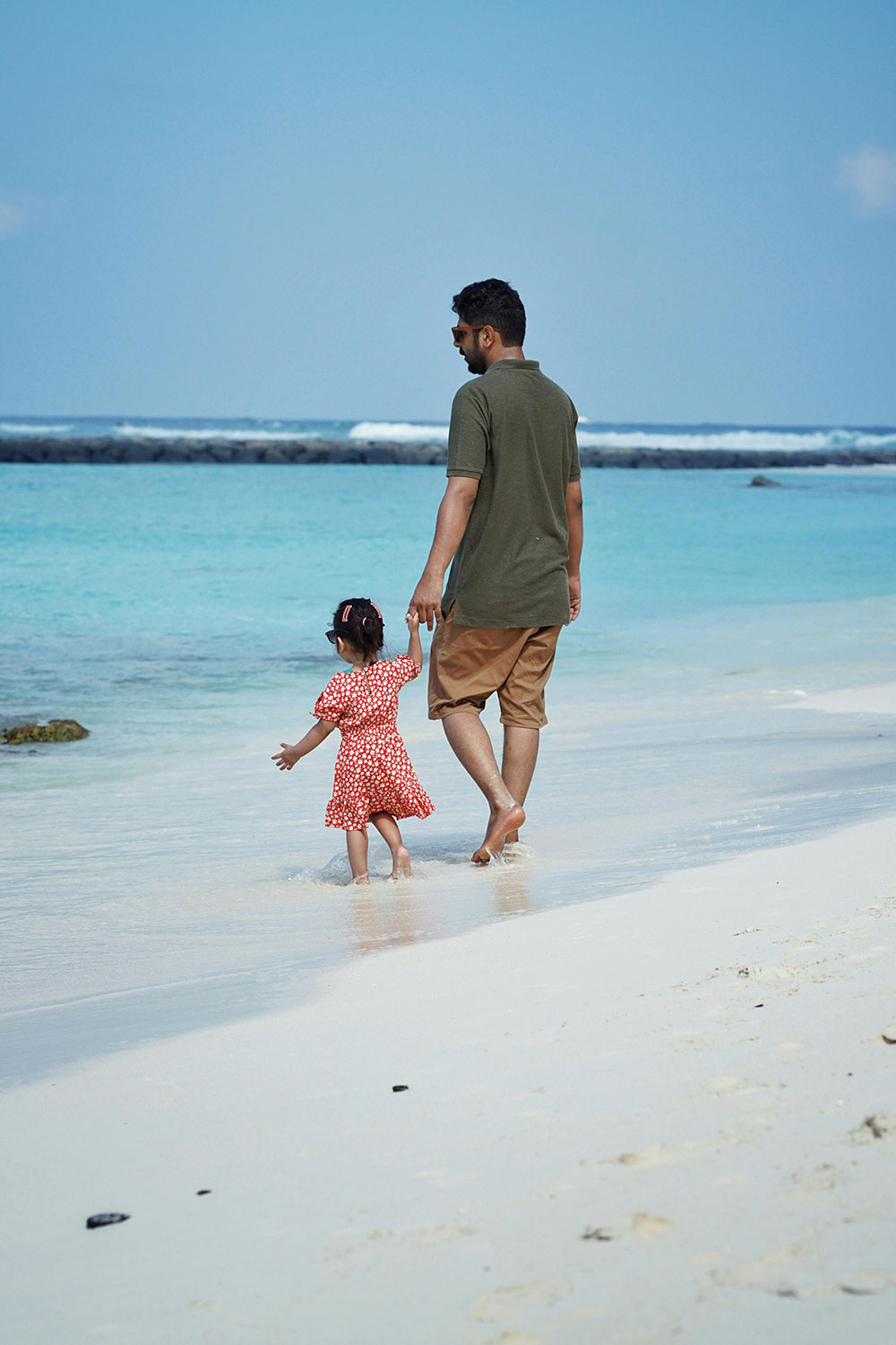 father wearing a casual polo shirt and shorts walking with a young child in a dress on a tropical beach