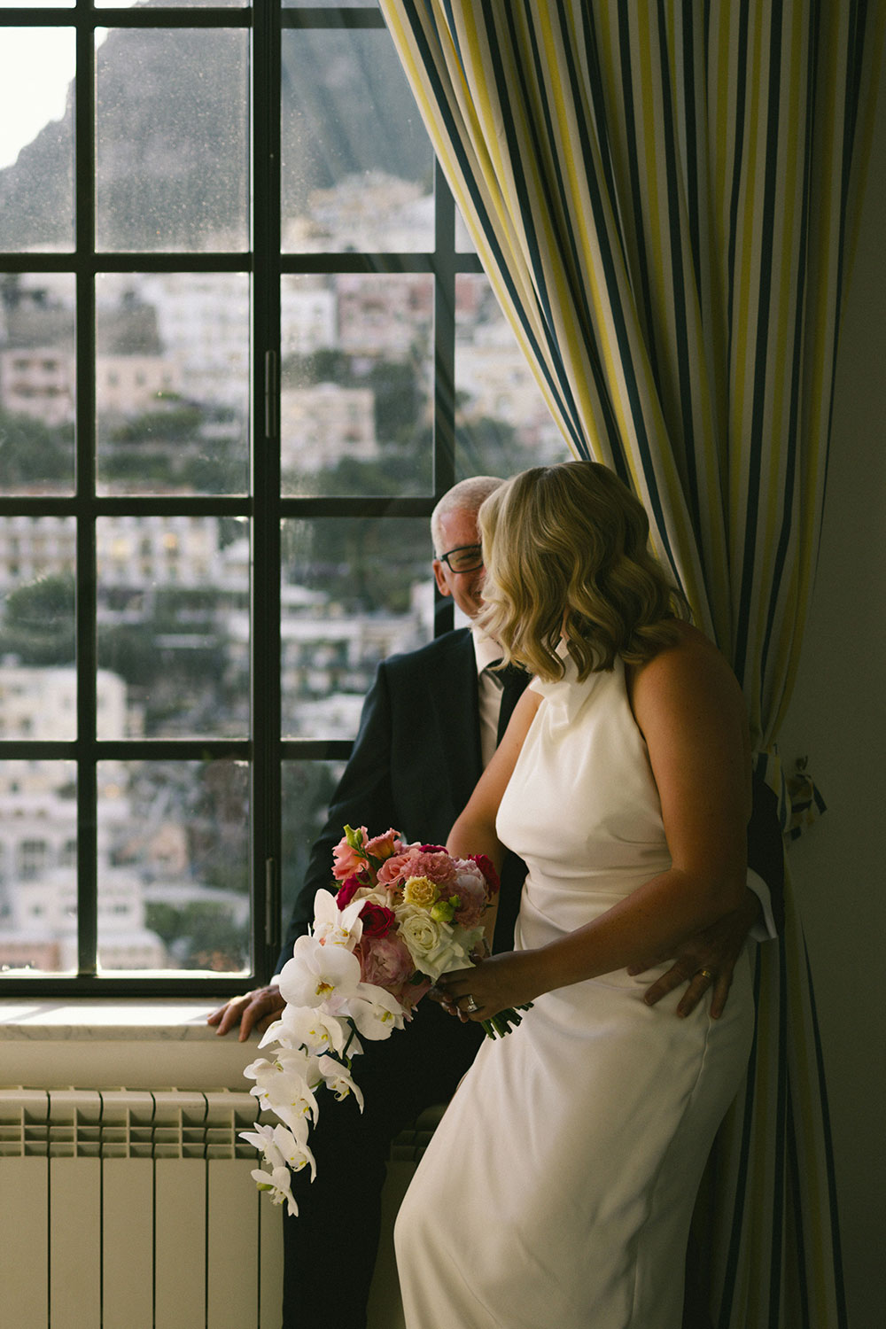 couple on their wedding day look out a window with a view of the Amalfi Coast in Italy