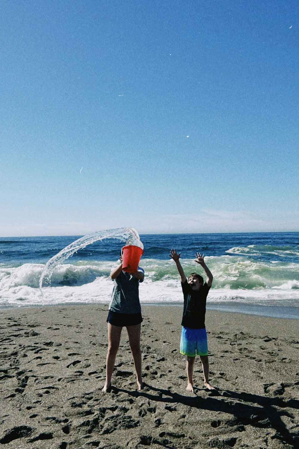 two kids playing on the beach with a water bucket